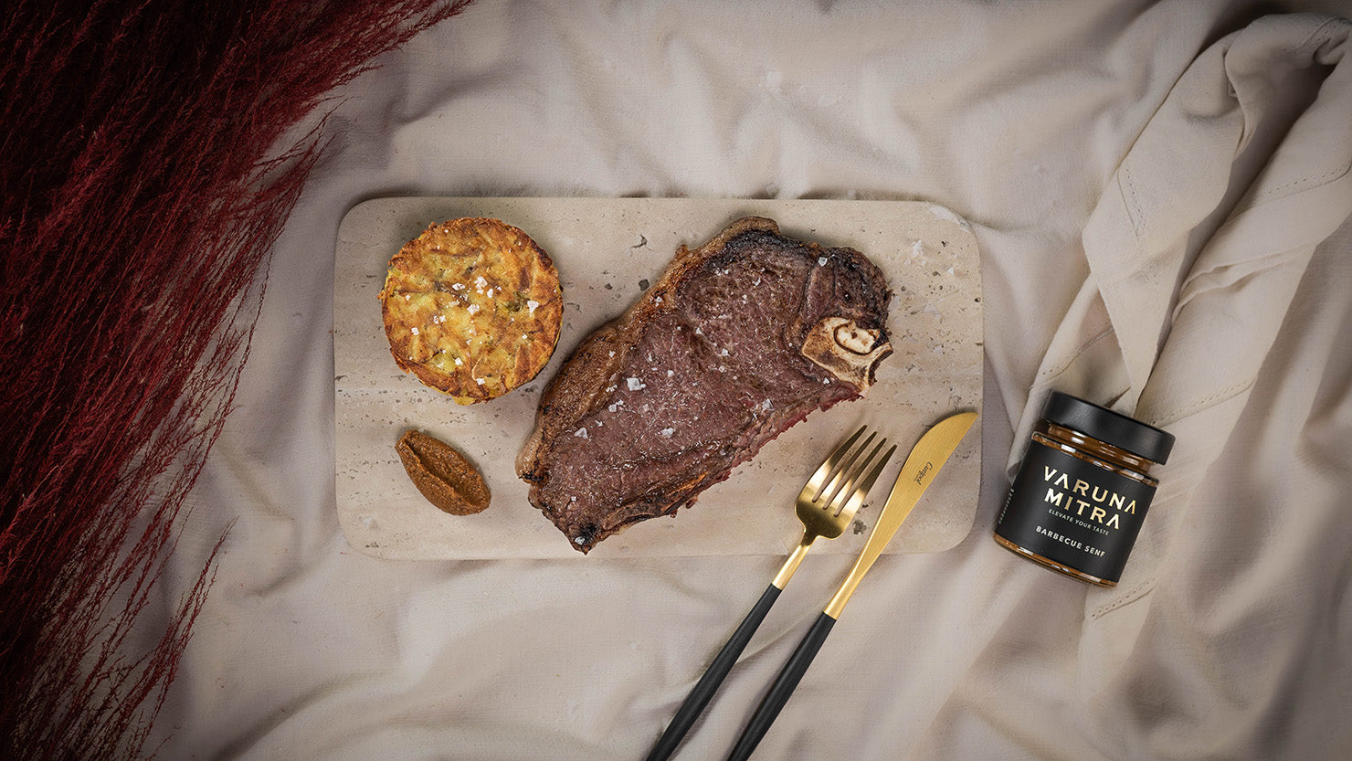 Varuna Mitra Barbecue Senf Glas neben goldenem Besteck, Rindersteak und einem Kartoffelpuffer auf einer Marmorplatte.
