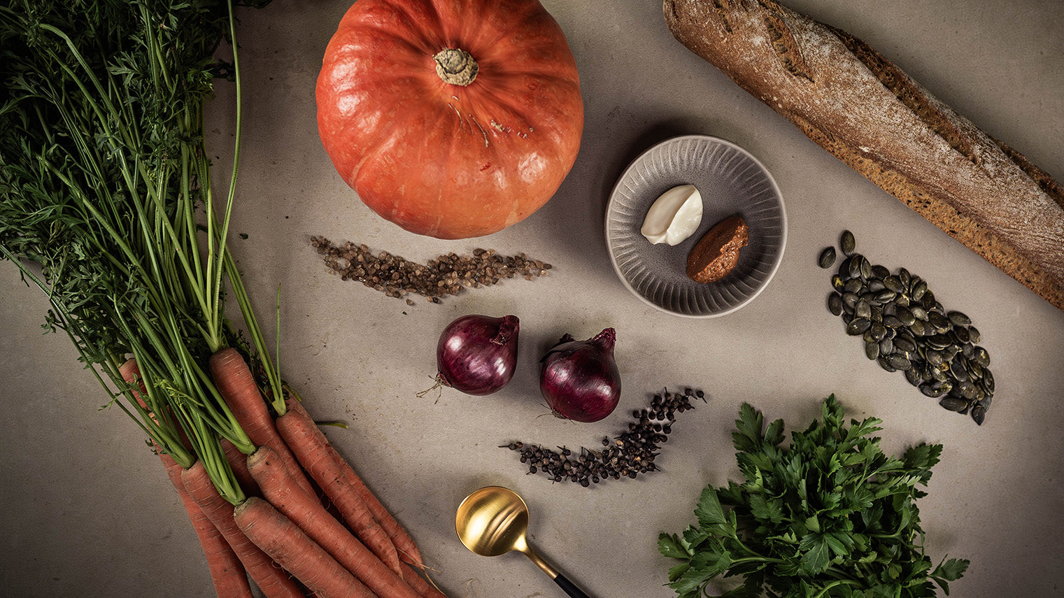 Zutaten wie Kürbis, Karotten, Zwiebeln, Kürbiskerne, Senf, Öl und Salz für eine Kürbiscreme Suppe auf neutralem Stein.