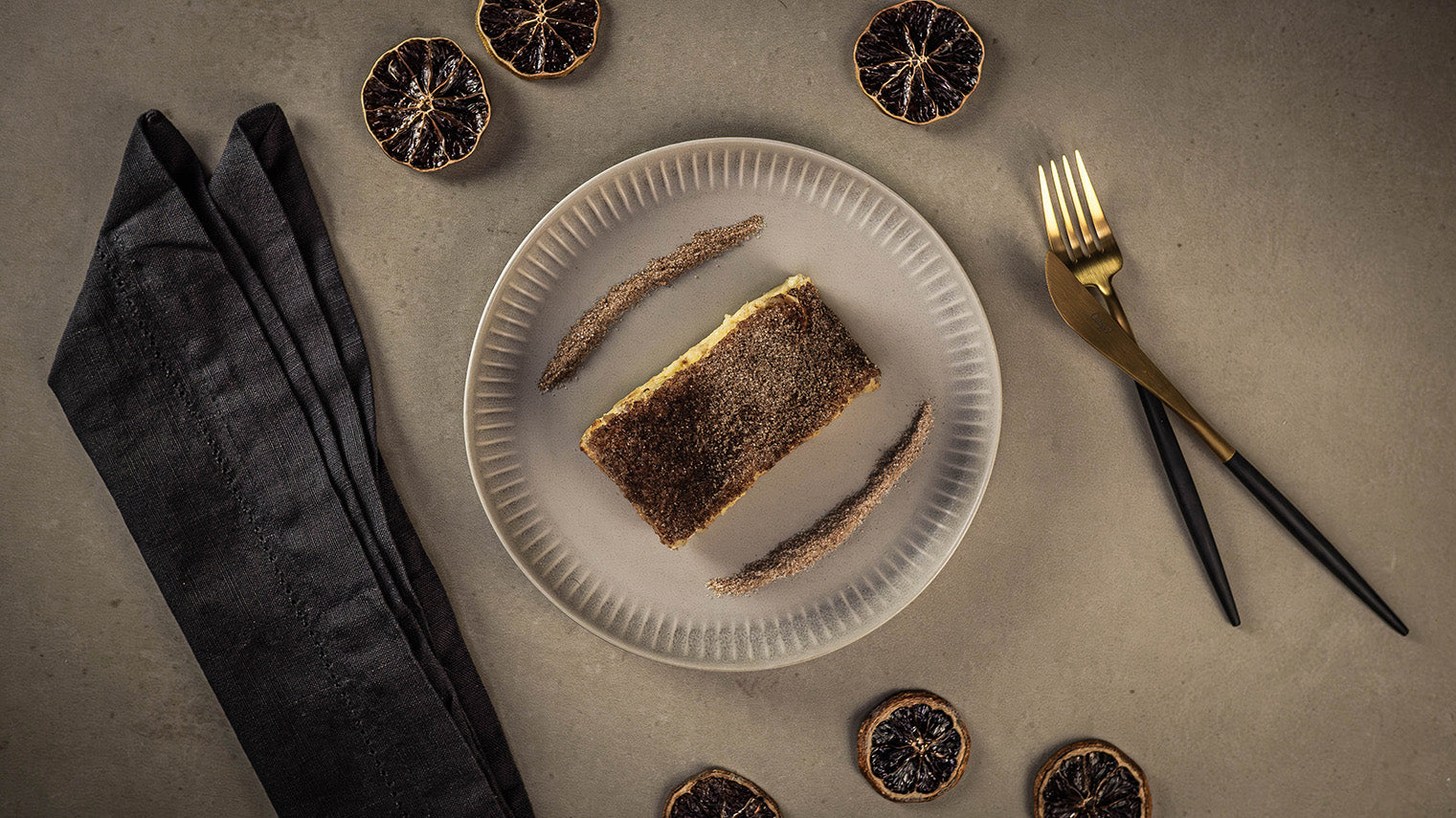 Rezept Zitronen Churro Cheesecake mit Zitronenöl auf einem Teller, neben getrockneten Zitronenscheiben, schwarzer Serviette und goldenem Besteck.