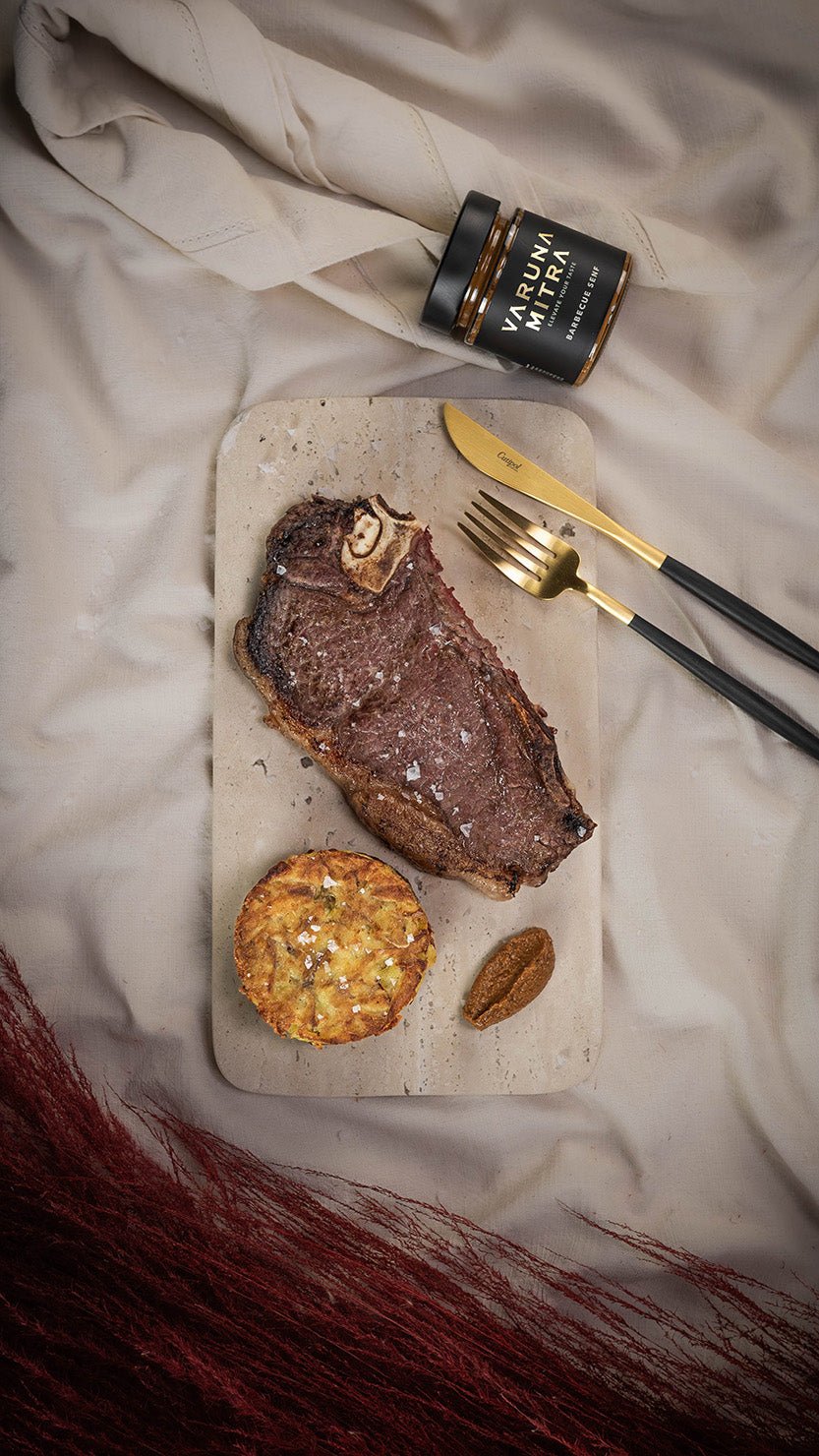 Varuna Mitra Barbecue Senf Glas neben goldenem Besteck, Rindersteak und einem Kartoffelpuffer auf einer Marmorplatte.