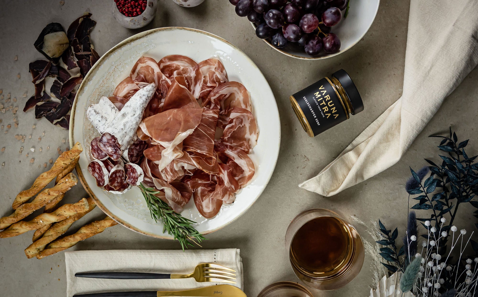 Tapas Teller mit scharfem Senf, Salami, Schinken und einer Schüssel roter Trauben, Brotstangen und Schinusbeeren Pfeffer.