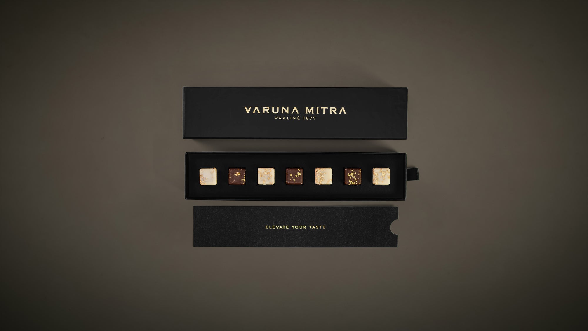 Varuna Mitra 7er schwarze Pralinenbox gefüllt mit 4 Zitrone-Pistazie Pralinen und 3 Kaffee-Karamell Pralinen. Abgebildet auf grün-grauen Hintergrund.